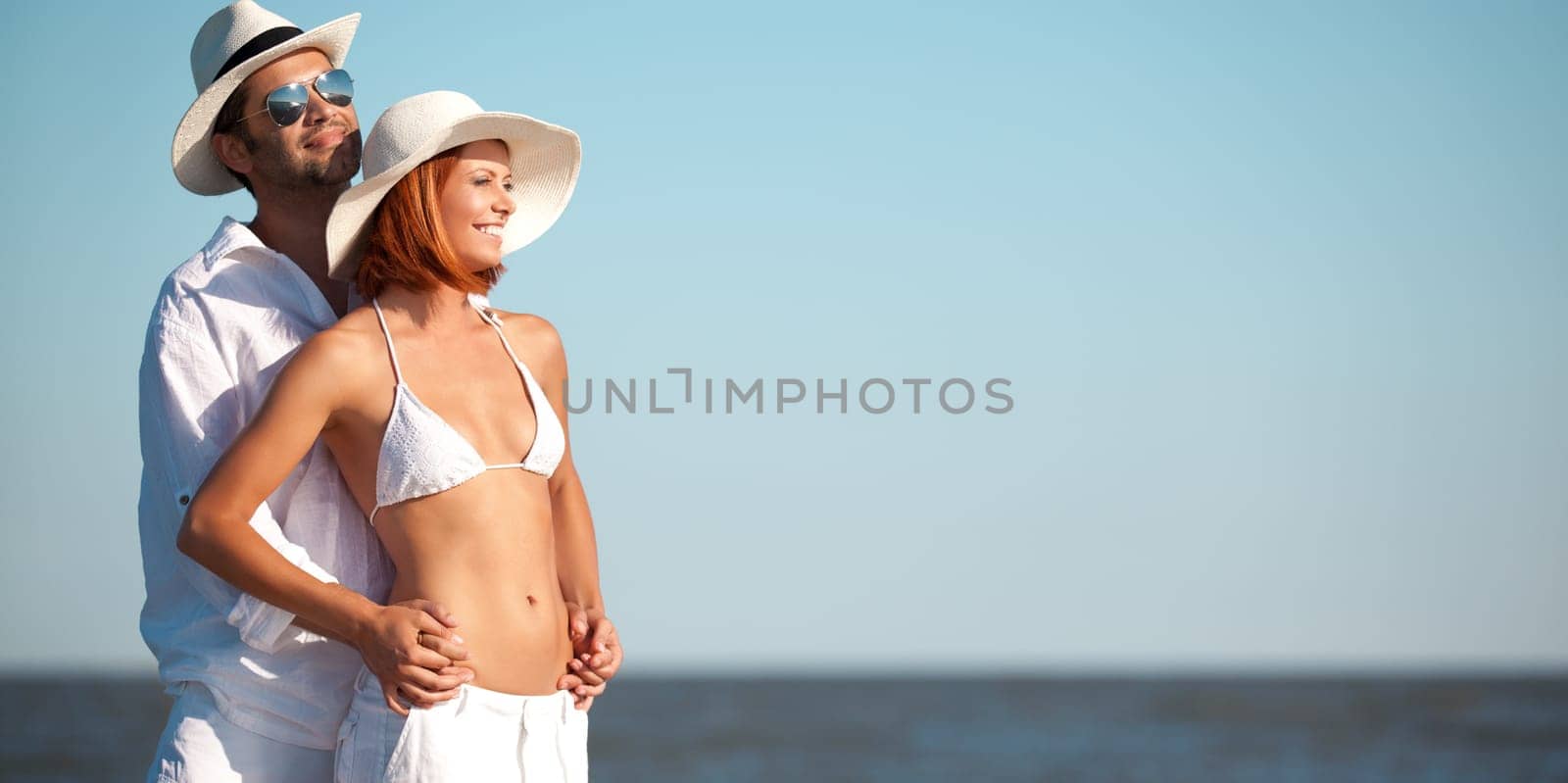 happy young couple, dressed in white, standing on a beach, smiling, holding each other