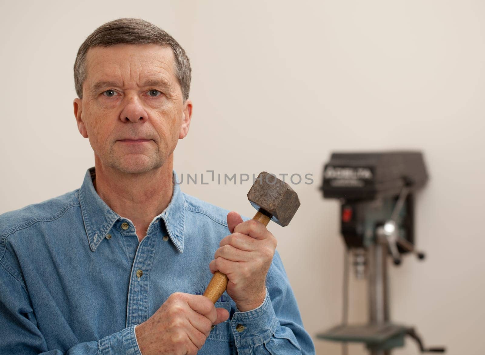 Senior male in a home workshop facing the camera and holding a large lump hammer