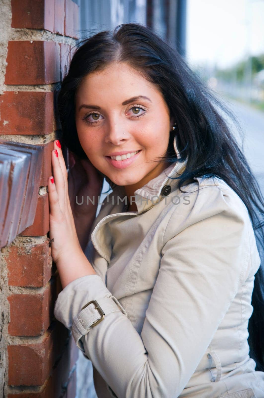 Upper body shot of a young,black-haired woman with long hair in a trench coat leaning against a brick wall and looking into the camera