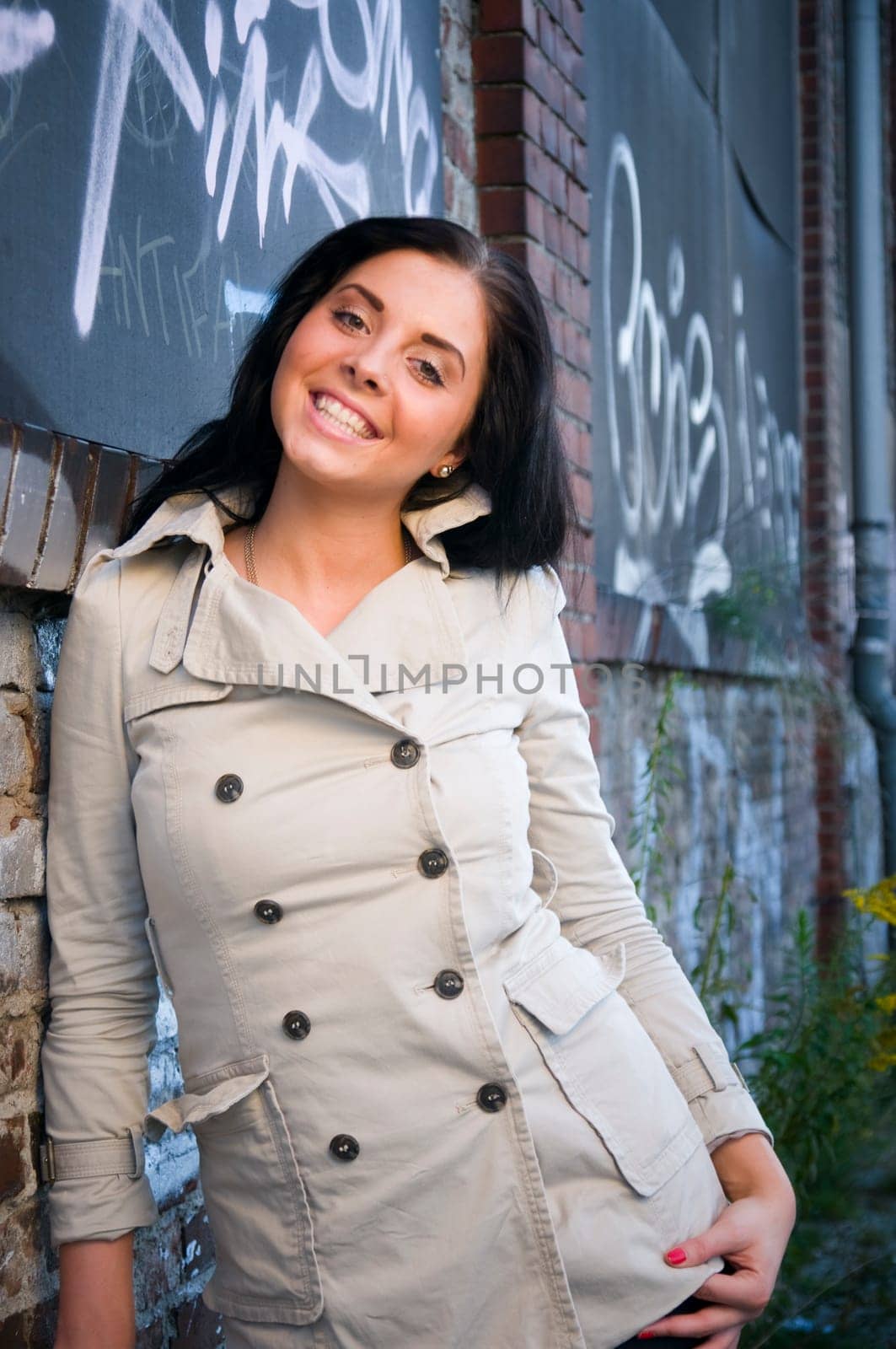 Upper body shot of a young,black-haired woman with long hair in a trench coat leaning against a brick wall and looking into the camera