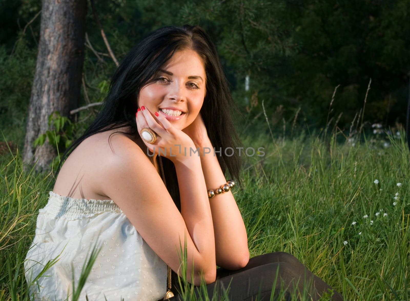 Young black-haired woman sitting in the grass resting elbows on knees and smiling at camera