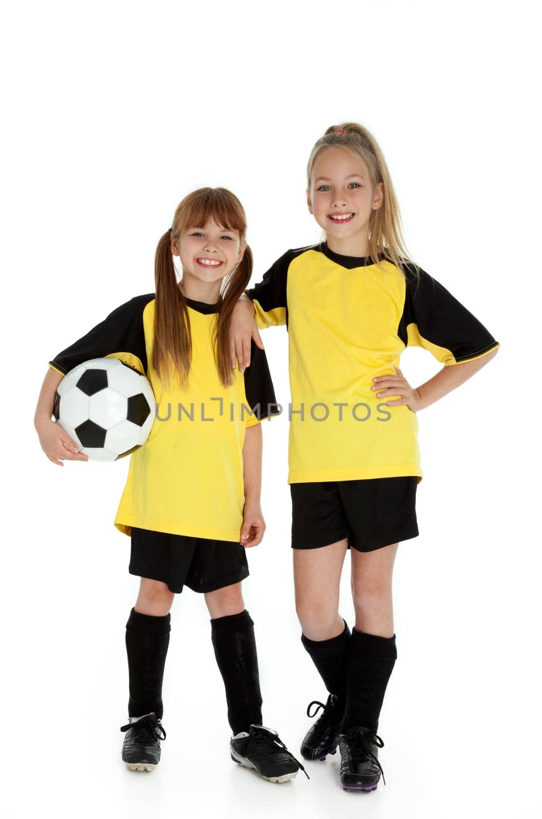 Full length front view of two young girls in soccer uniforms, holding soccer ball on white.