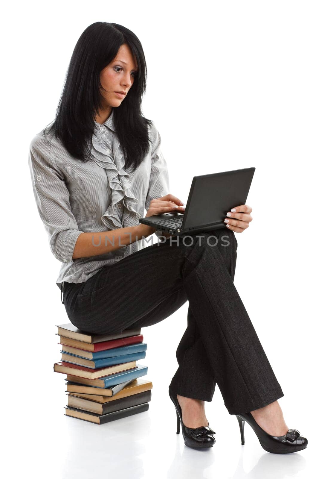 The attractive student sits on a pile of books with the laptop on a white background.