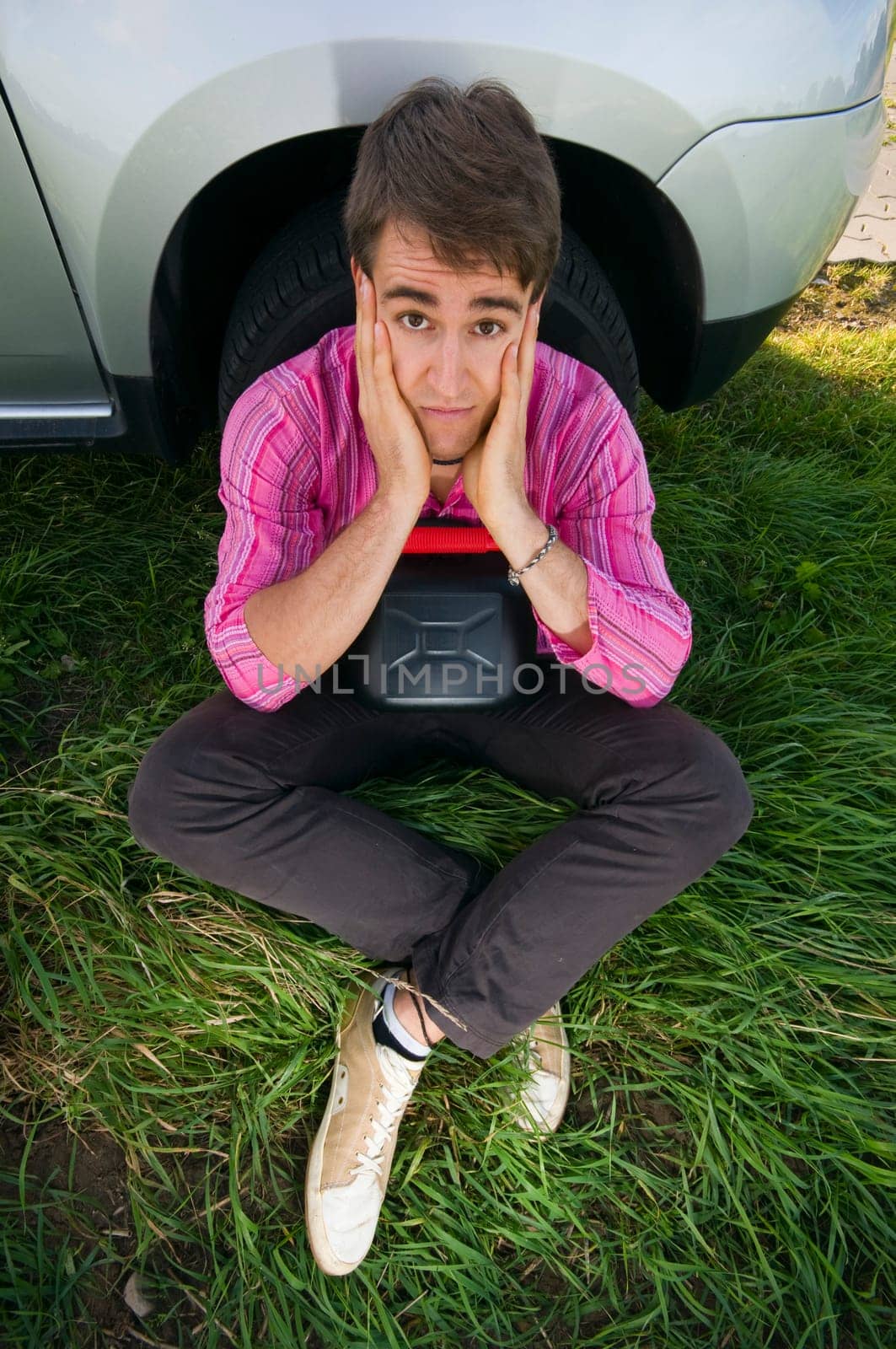 Young man sitting at the front wheel of a vehicle with a black gasoline canister and looking helplessly at the camera