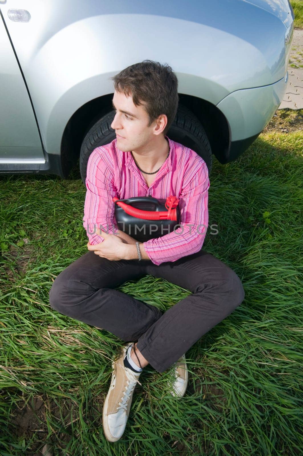 Young man sitting at the front wheel of a vehicle with a black gasoline canister and looking to the side helplessly
