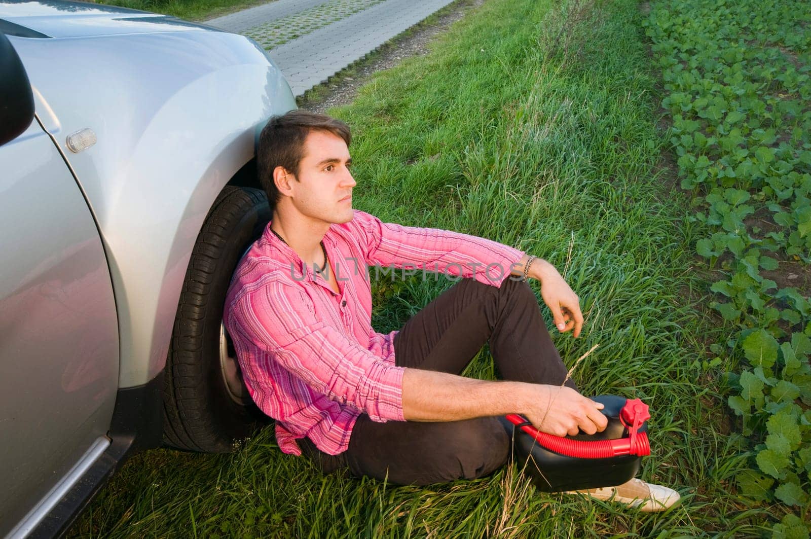 young man sits on the side of the road in the grass at the front wheel of a silver-gray car and handles a black gasoline canister