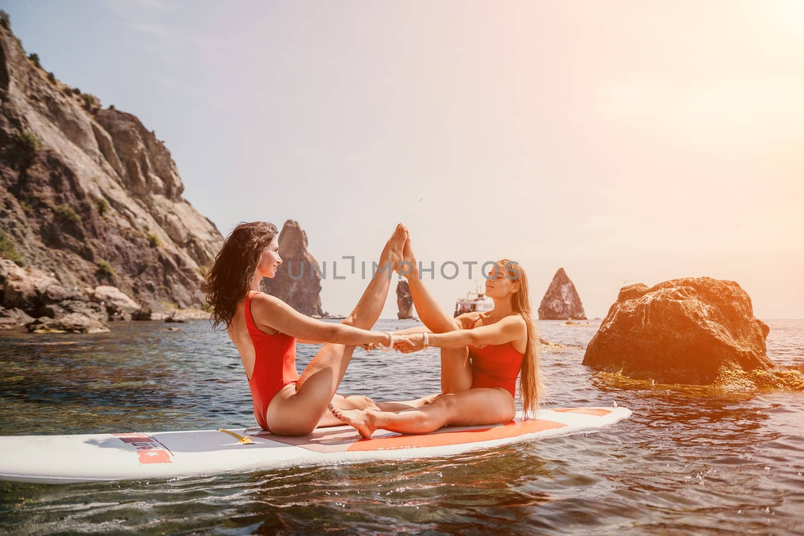 Woman sup yoga. Two happy sporty women practising yoga pilates on paddle sup surfboard. Female stretching doing workout on sea water. Modern individual female hipster outdoor summer sport activity. by panophotograph