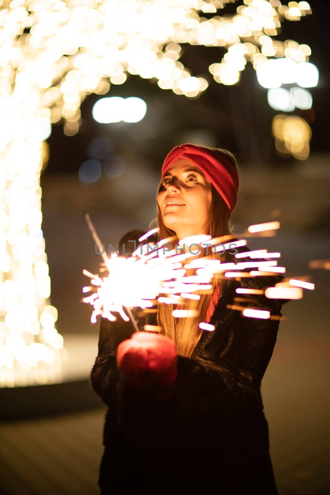 Woman holding sparkler night while celebrating Christmas outside. Dressed in a fur coat and a red headband. Blurred christmas decorations in the background. Selective focus.