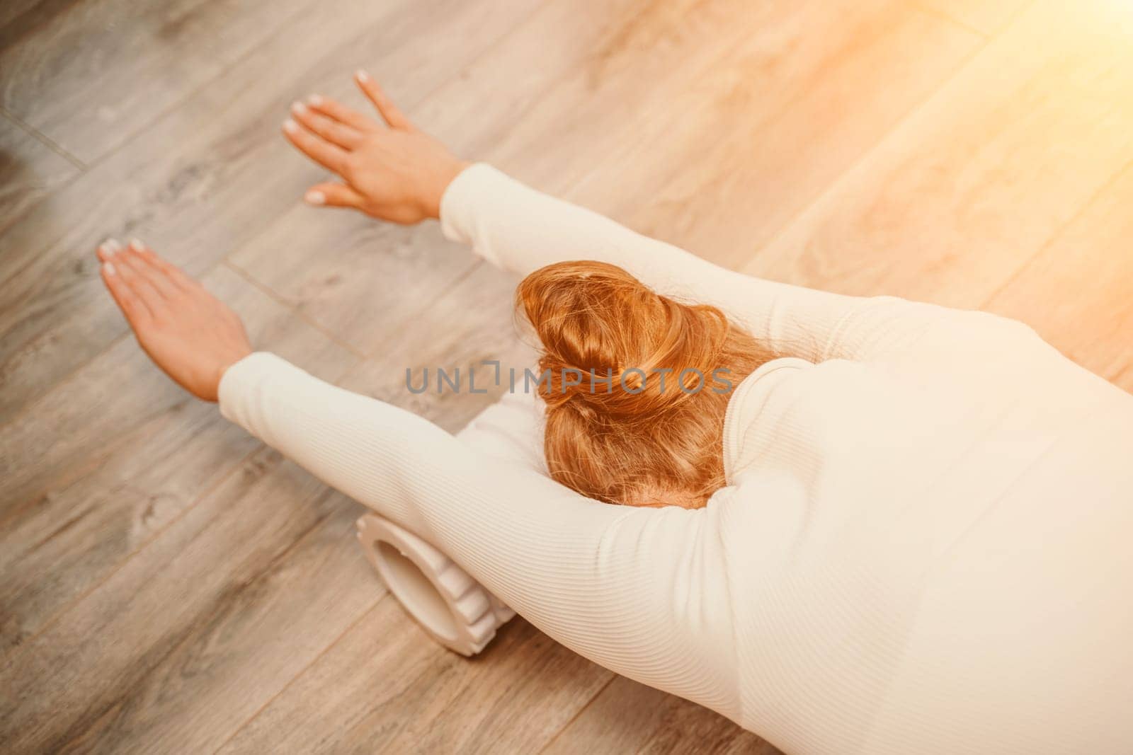 Adult athletic woman, in white bodysuit, performing fascia exercises on the floor - caucasian woman using a massage foam roller - a tool to relieve tension in the back and relieve muscle pain - the concept of physiotherapy and stretching training by Matiunina