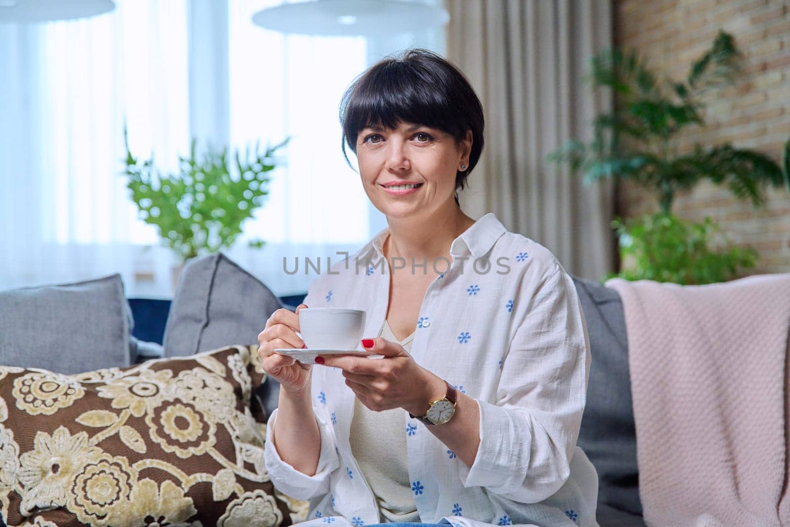 Smiling mature beautiful woman relaxing at home with cup of coffee tea. 50s positive female sitting on couch in living room, looking at camera. Age people, lifestyle, beauty, health concept