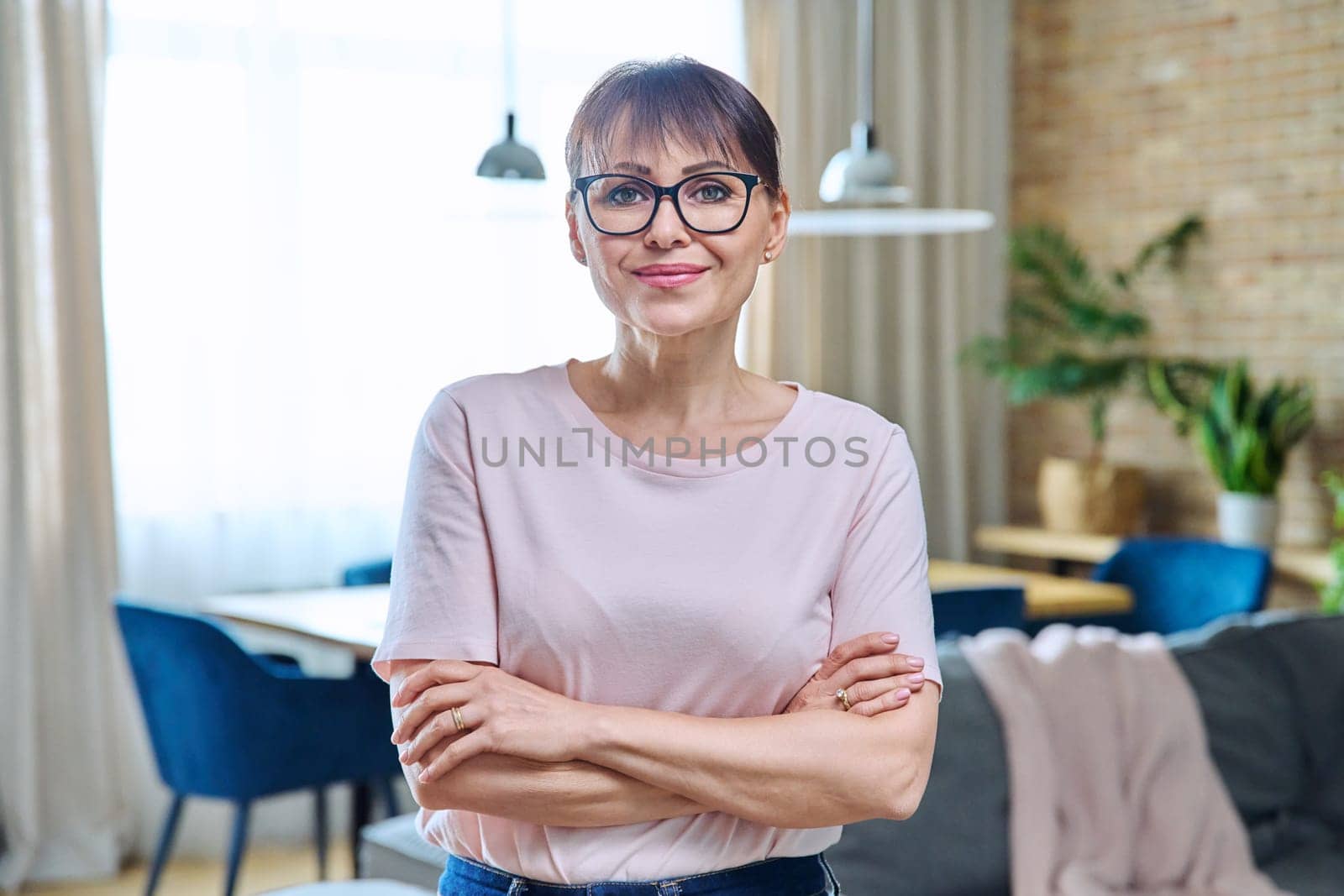 Portrait of positive confident middle aged woman at home. Smiling successful female with crossed arms looking at camera, living room interior background