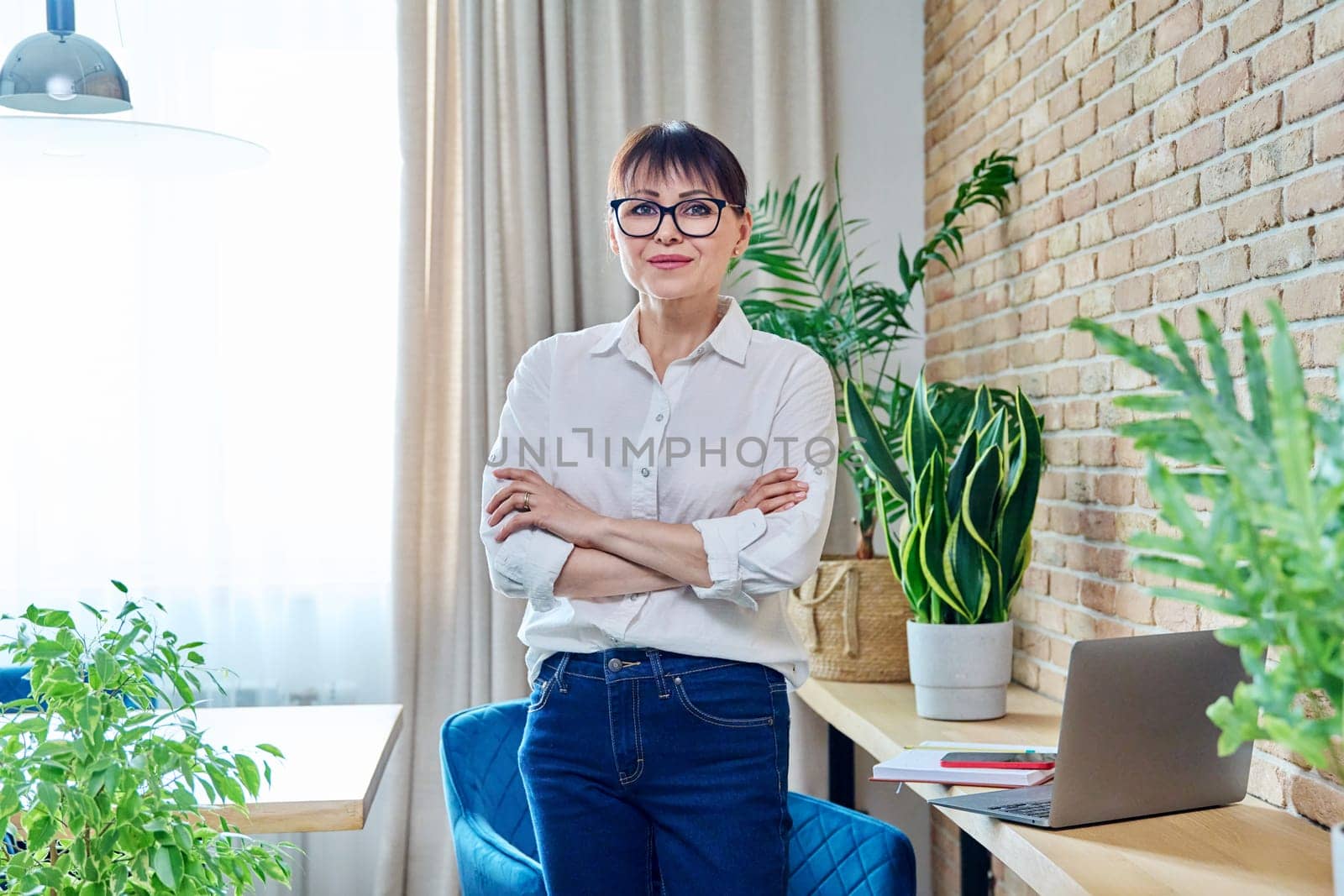 Portrait of positive confident middle aged woman at home. Smiling successful female with crossed arms looking at camera, living room interior background