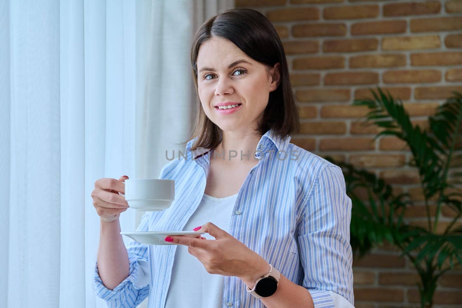 Young smiling woman with cup of morning coffee at home near window. Attractive 30 year old female looking at camera. Lifestyle, beauty, people concept