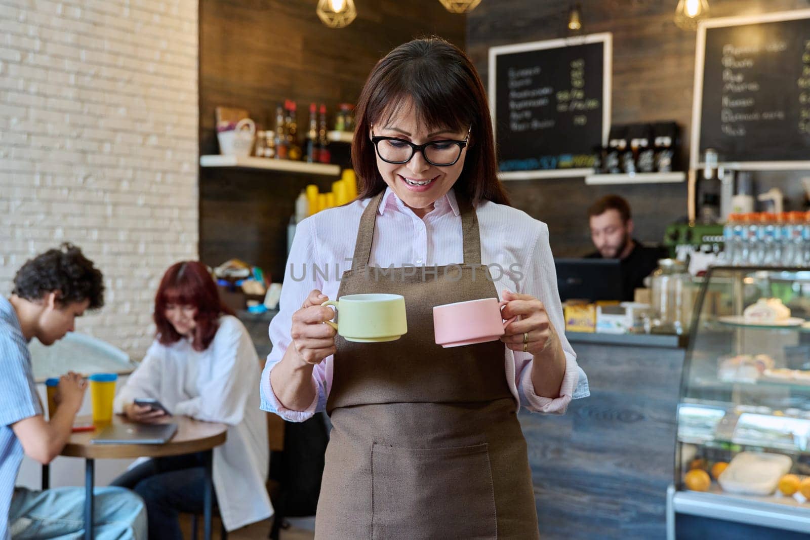 Portrait of smiling female coffee shop worker holding two cups of coffee in her hands. Woman in apron looking at camera inside cafe hall. Small business, food service occupation, staff, work concept