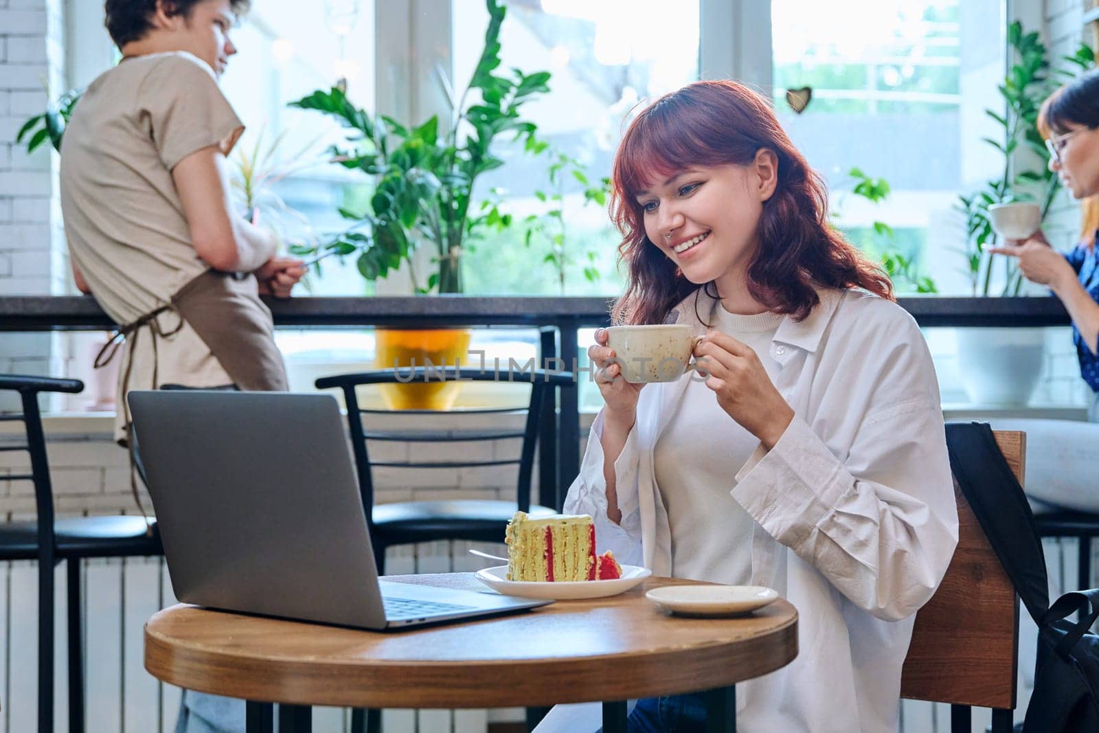 Female college student with laptop in cafe at table with cup of coffee and piece of cake. Internet online technology for leisure communication blogging learning chat, youth lifestyle concept