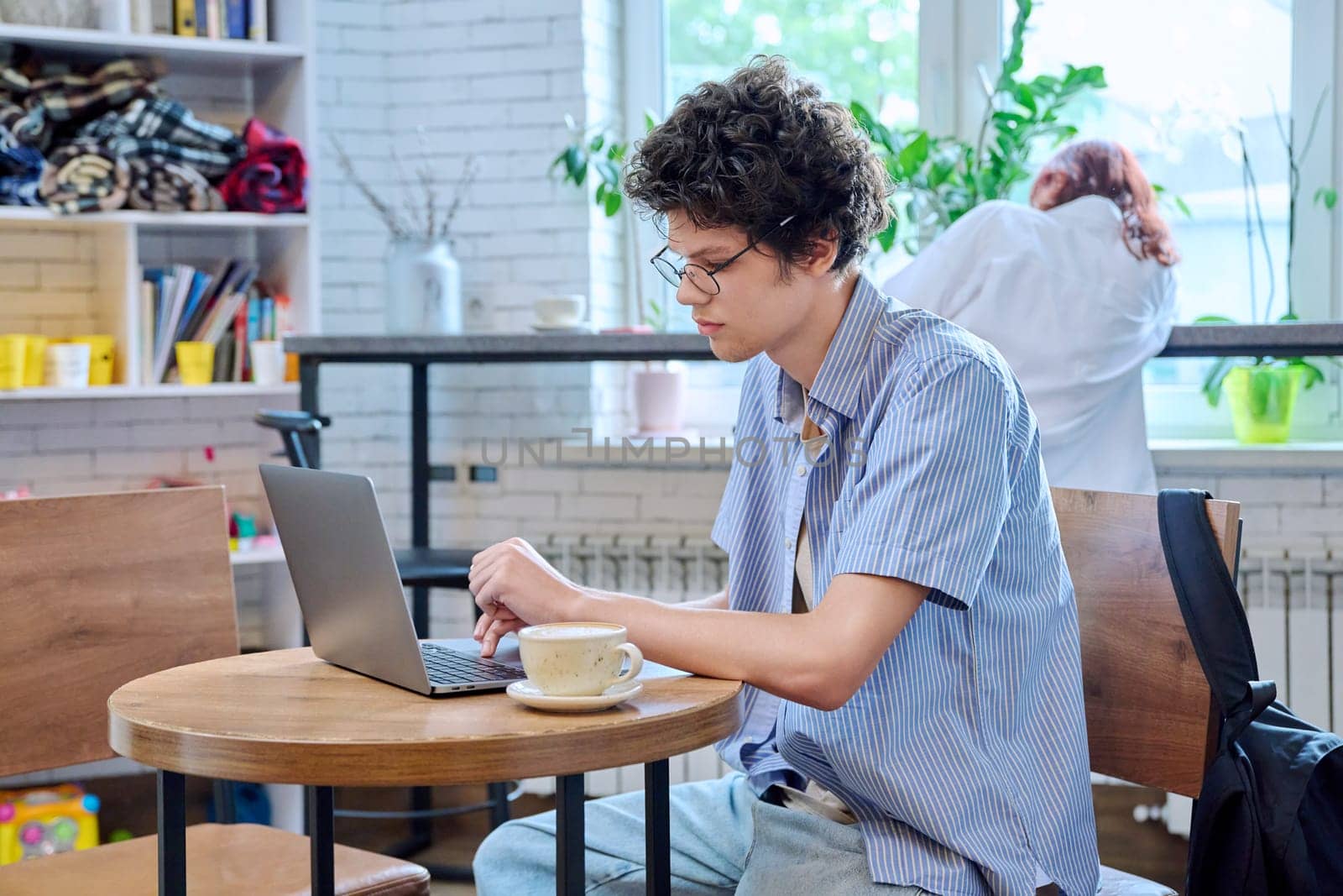 Handsome young curly guy college student typing on laptop sitting at table at coffee shop with cup of coffee. Internet online technology for leisure communication learning, chat, blog, youth lifestyle