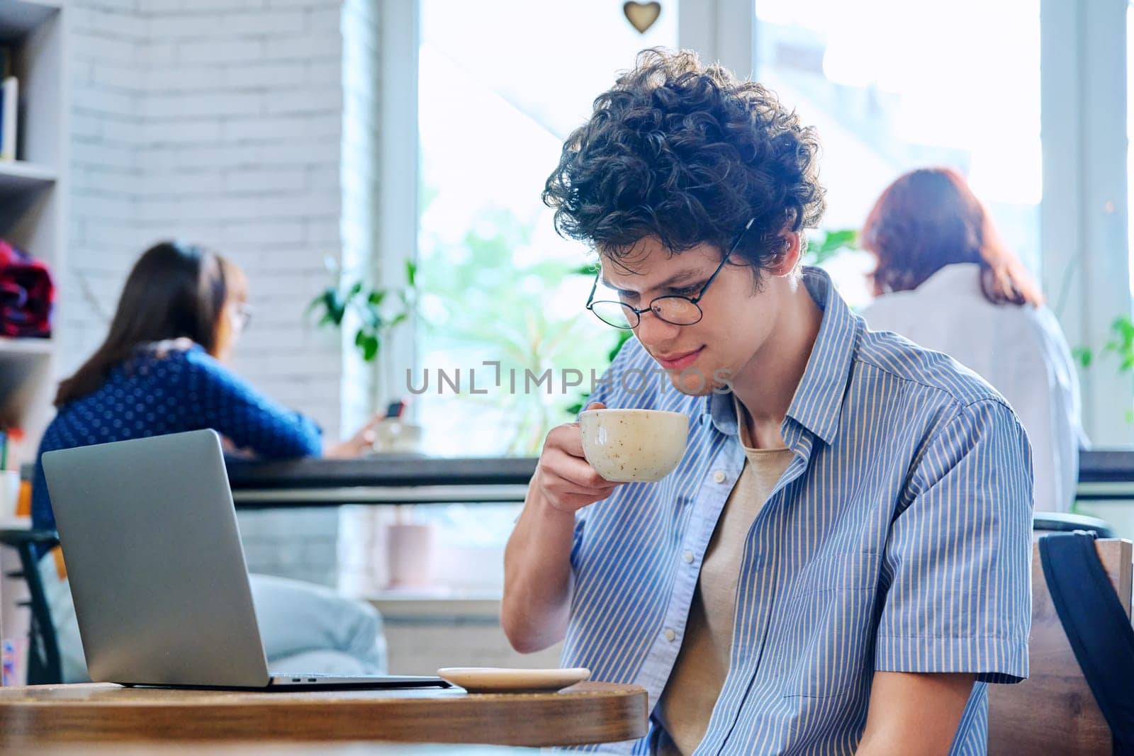 Handsome young curly-haired guy drinking cup of coffee in coffee shop. Young male college student in glasses enjoying cappuccino. Coffee culture, lifestyle, youth, people concept