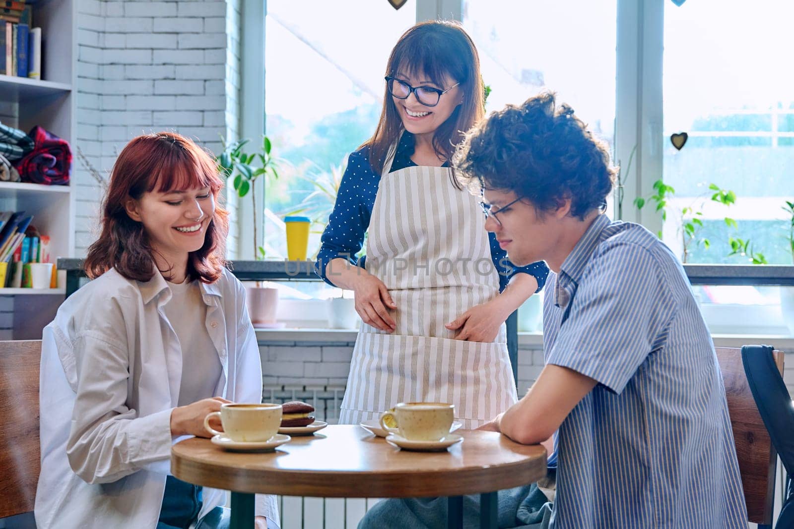 Woman in an apron with cup of coffee and plate of cake serving young people in cafeteria by VH-studio