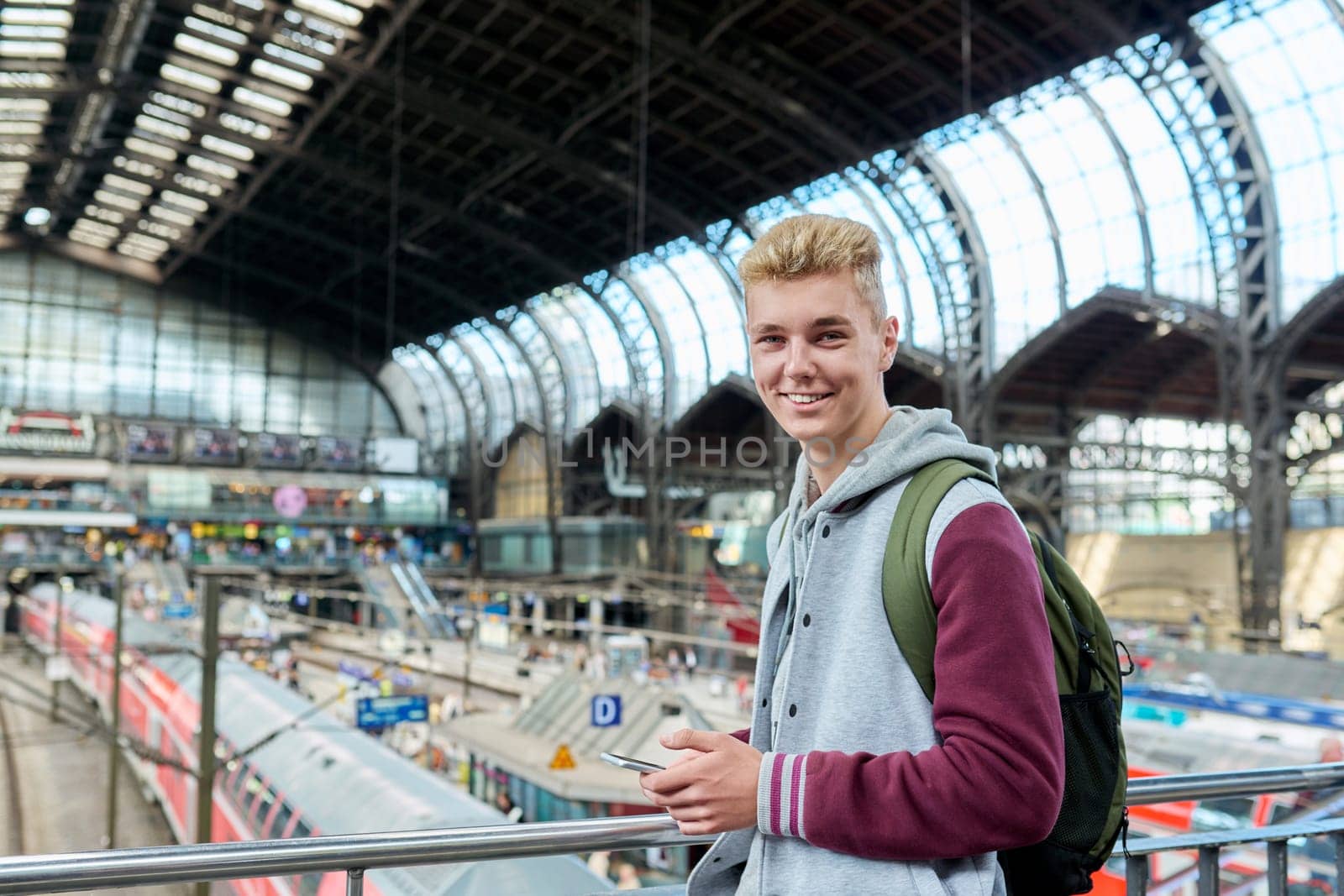 Portrait of smiling young guy 18, 19 years old with backpack smartphone looking at camera at commuter train station. Youth, urban lifestyle, passenger rail transport
