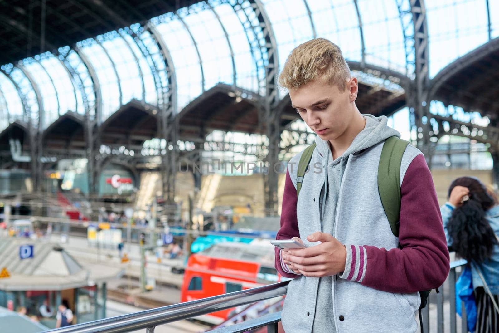 Young guy using mobile apps on smartphone standing at railway station by VH-studio