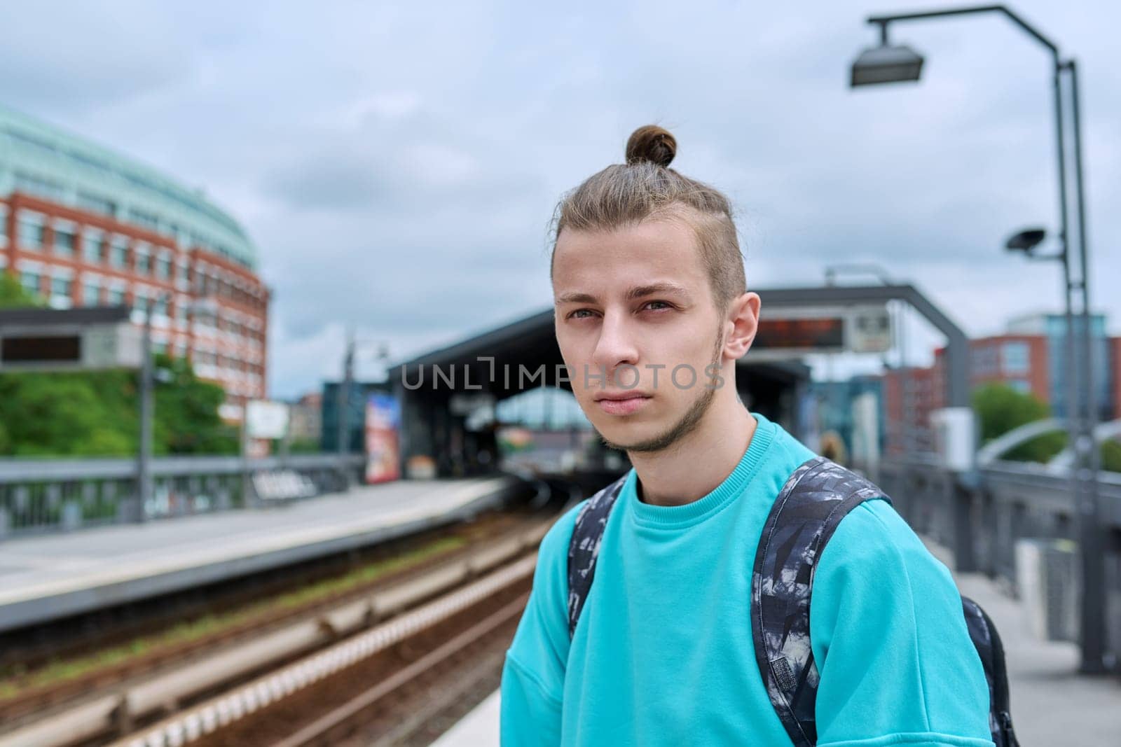 Young male waiting for electric train at city railway platform station. Guy serious student hipster 19, 20 years old with backpack. Urban transport, commuter train, passenger transportation, youth