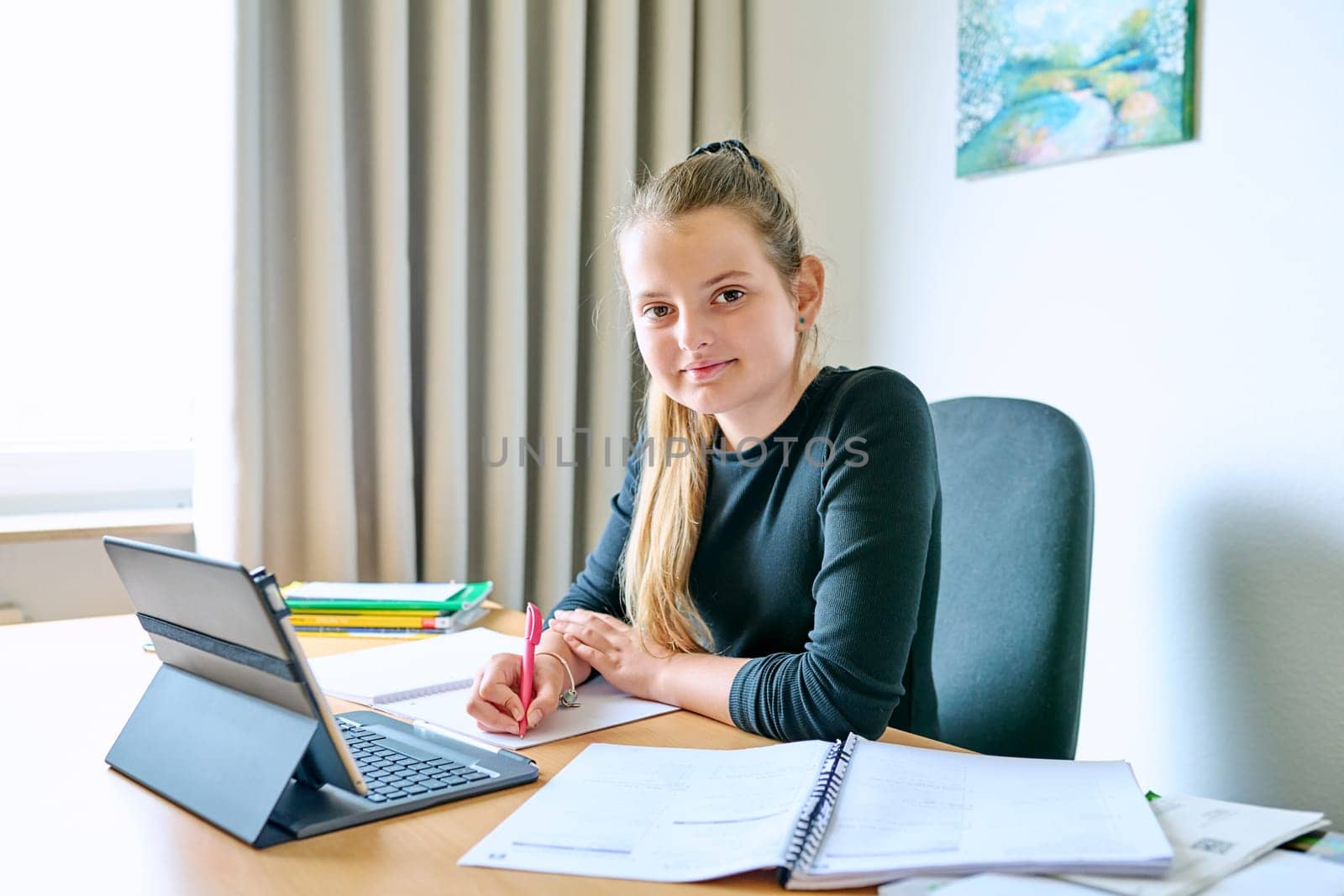 Portrait of smiling child girl sitting at desk at home with digital tablet textbooks, looking at camera. Education, knowledge, school, childhood concept