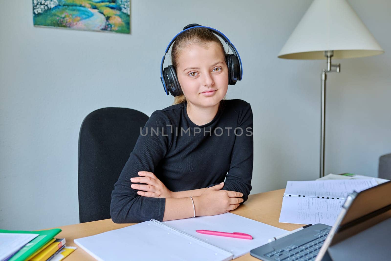 Portrait of smiling child girl in headphones sitting at desk at home with digital tablet textbooks, looking at camera. Education, knowledge, school, childhood concept