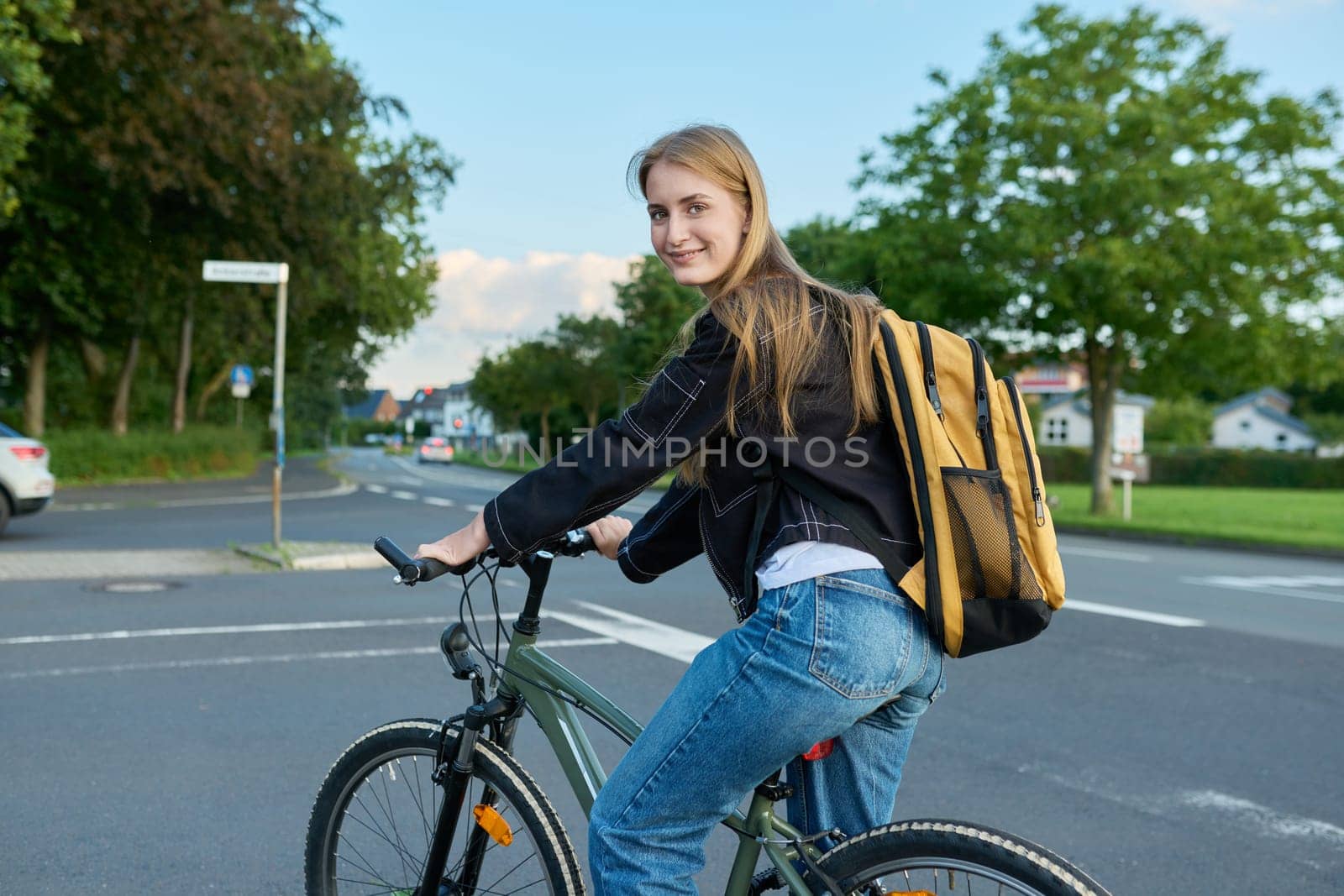 Portrait of teenage student girl with backpack on bicycle. Smiling teen female 16, 17 years old looking at camera outdoors on road. High school, lifestyle, adolescence, youth concept