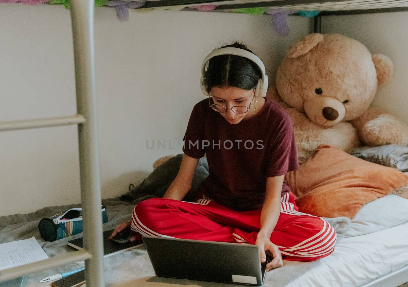 One young beautiful Caucasian teenage girl with white headphones and sportswear sits on a bunk bed with a large toy bear and teaches homework at the computer, side view close-up.