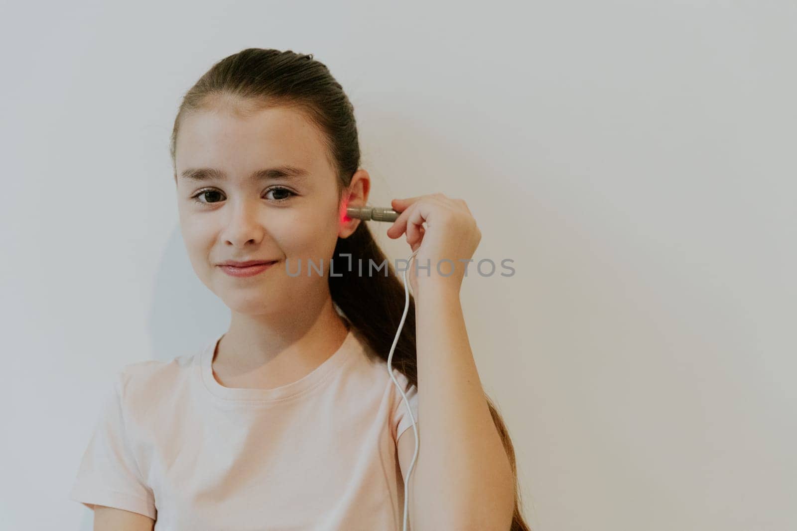 One beautiful Caucasian brunette girl with collected hair and in a pink T-shirt treats her ear with an infrared light device, standing on the left near a white wall with copy space on the right, side view close-up.