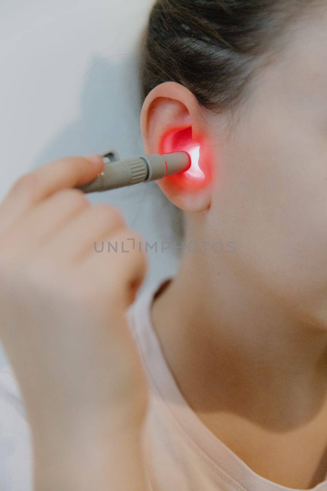 One beautiful Caucasian brunette girl with a happy smile, collected hair and in a pink T-shirt treats her right ear with an infrared light device, sitting on a bed at home near a white wall, side view very close-up.