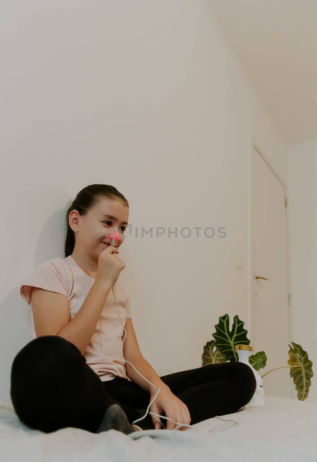 One beautiful Caucasian brunette girl with collected hair and in a pink T-shirt treats the right nasal passage with an apparatus with infrared light, sitting on the bed, cross-legged, leaning on a white wall, close-up view from below.
