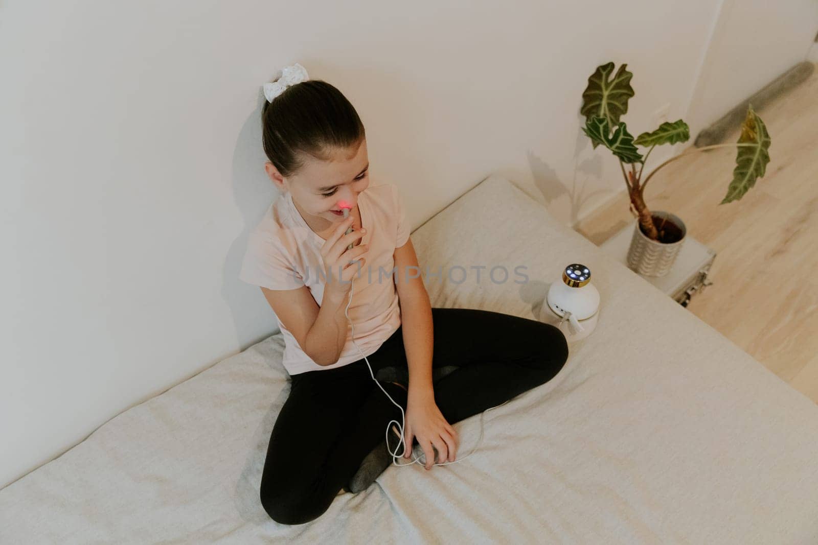 One beautiful Caucasian brunette girl with a happy smile, collected hair and in a pink T-shirt treats the right nasal passage with a device with infrared light, sitting on the bed, cross-legged, leaning on a white wall, close-up view from above.