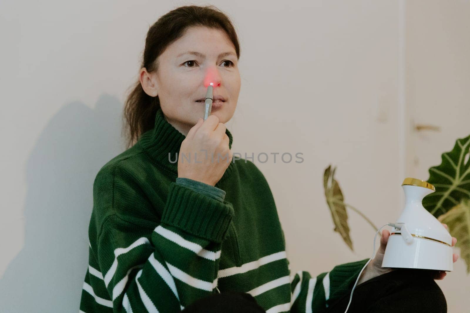 One beautiful Caucasian brunette girl with collected hair in a green sweater treats the right nasal passage with an apparatus with infrared light, sitting on a bed against a white wall, close-up view from below.