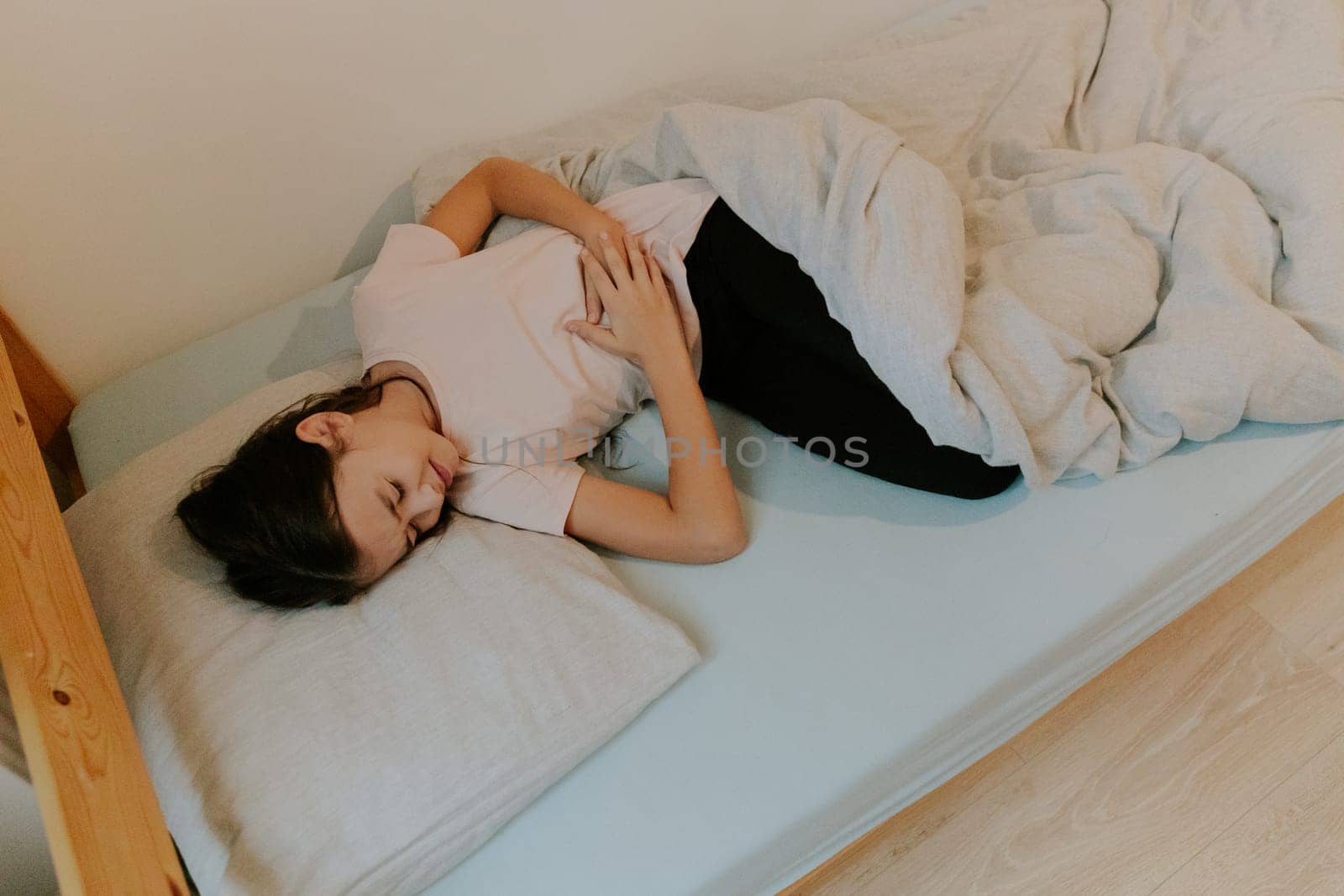One beautiful little Caucasian brunette girl with flowing long hair sleeps in bed, curled up in pain and holding her stomach with both hands, close-up side view.