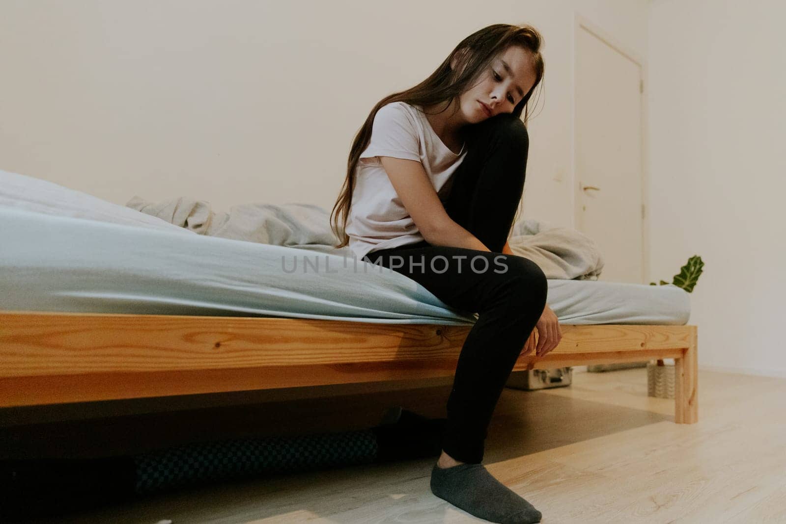 One beautiful little Caucasian brunette girl with a sad emotion on her face sits on the bed with one leg raised to her chin during the day in her room, close-up view from below.