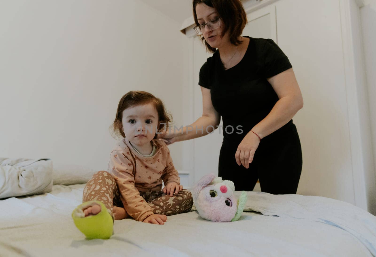 One Caucasian mother, talking, combing her baby daughter with a cast on her leg looking to the side and sitting on the bed in the morning in the room, side view close-up.