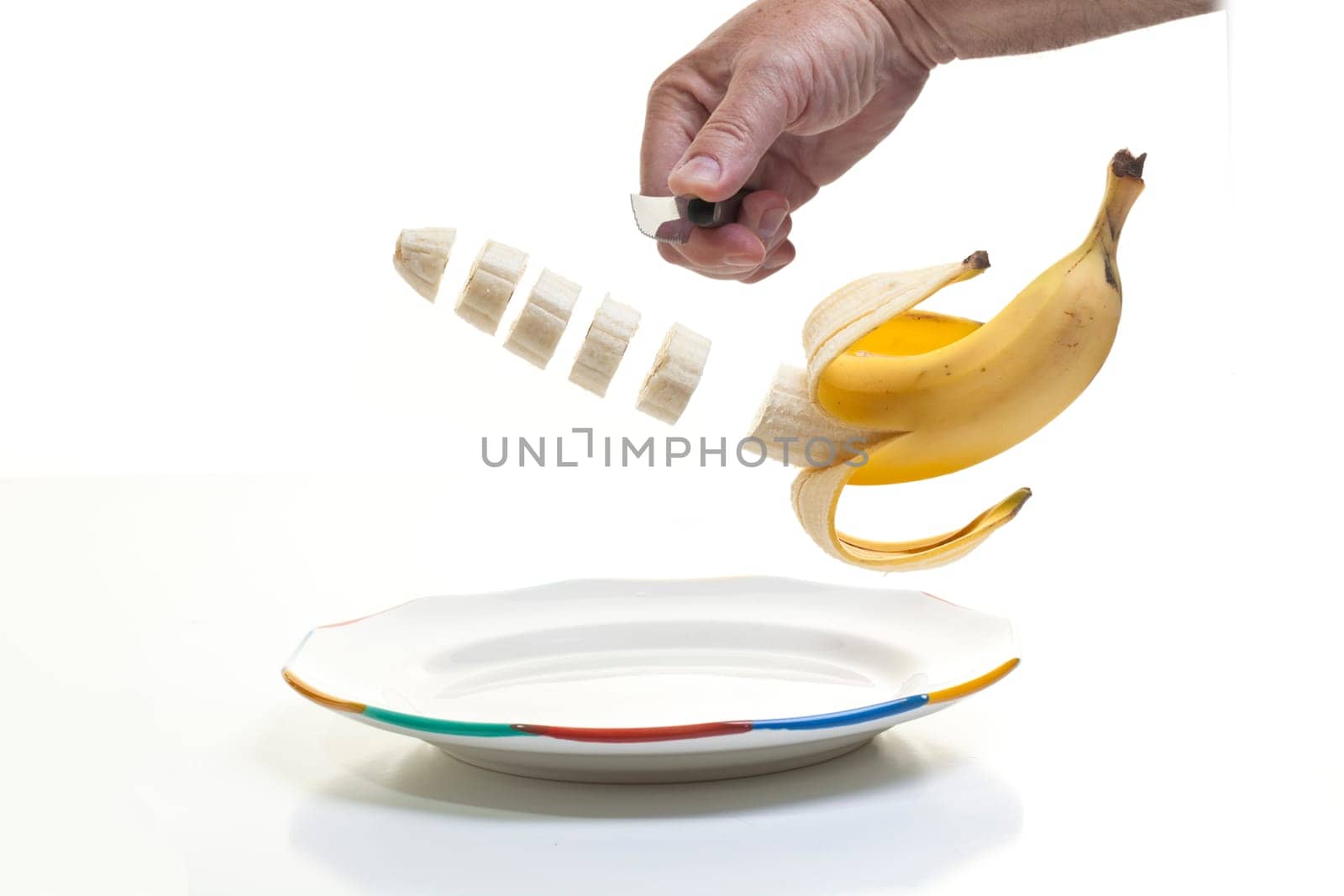 A human hand holding a knife and slicing a half peeled banana with the slices suspended midair over a plate