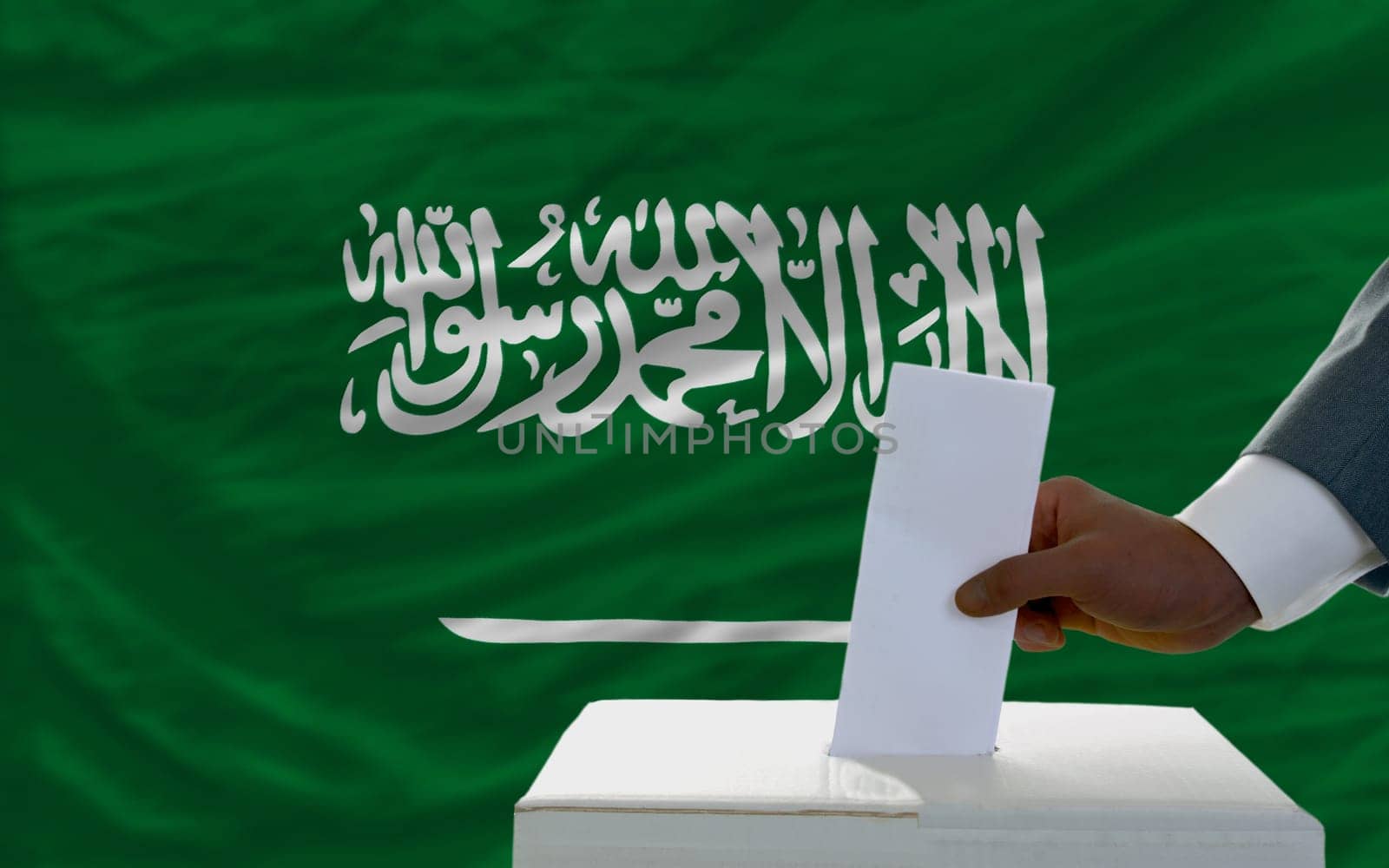 man putting ballot in a box during elections  in front of national flag of saudi arabia