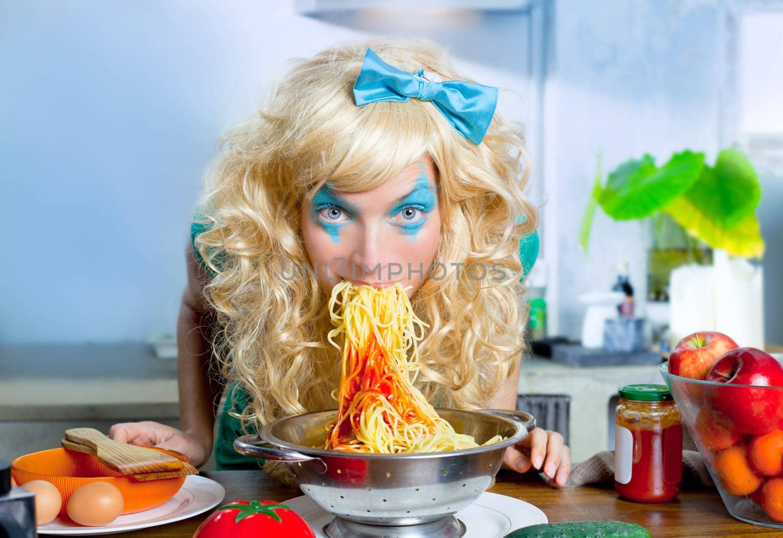 Blonde funny girl on kitchen eating pasta like crazy with blue makeup