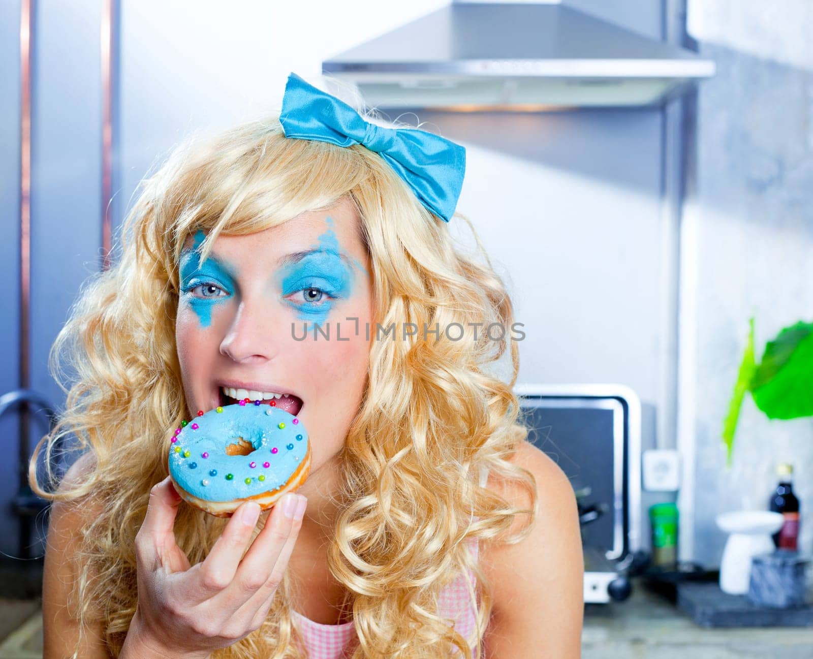 Blonde funny girl on kitchen eating blue donut with fashion makeup