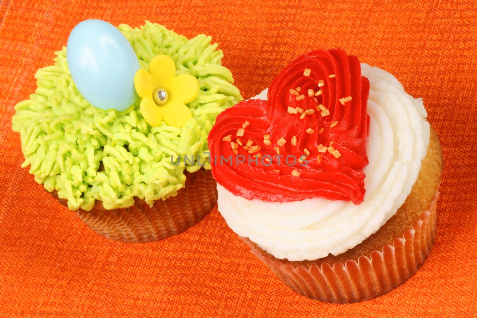 Two fancy holiday cupcakes over an orange background. A Valentine&#039 s day cupcake with a red buttercream heart on the right and an Easter cupcake decorated with a light blue hard sugar egg and a yellow flower on the left.