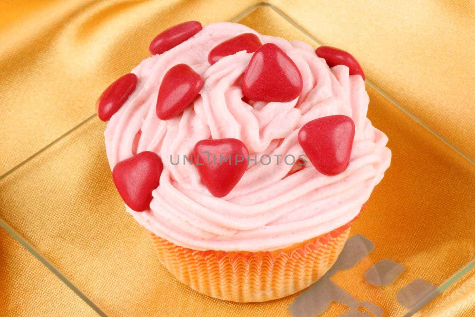 Fancy Valentine&#039 s Day cupcake decorated with small red sugar-coated chocolate hearts on a transparent glass dish, over a yellow background. Selective focus.