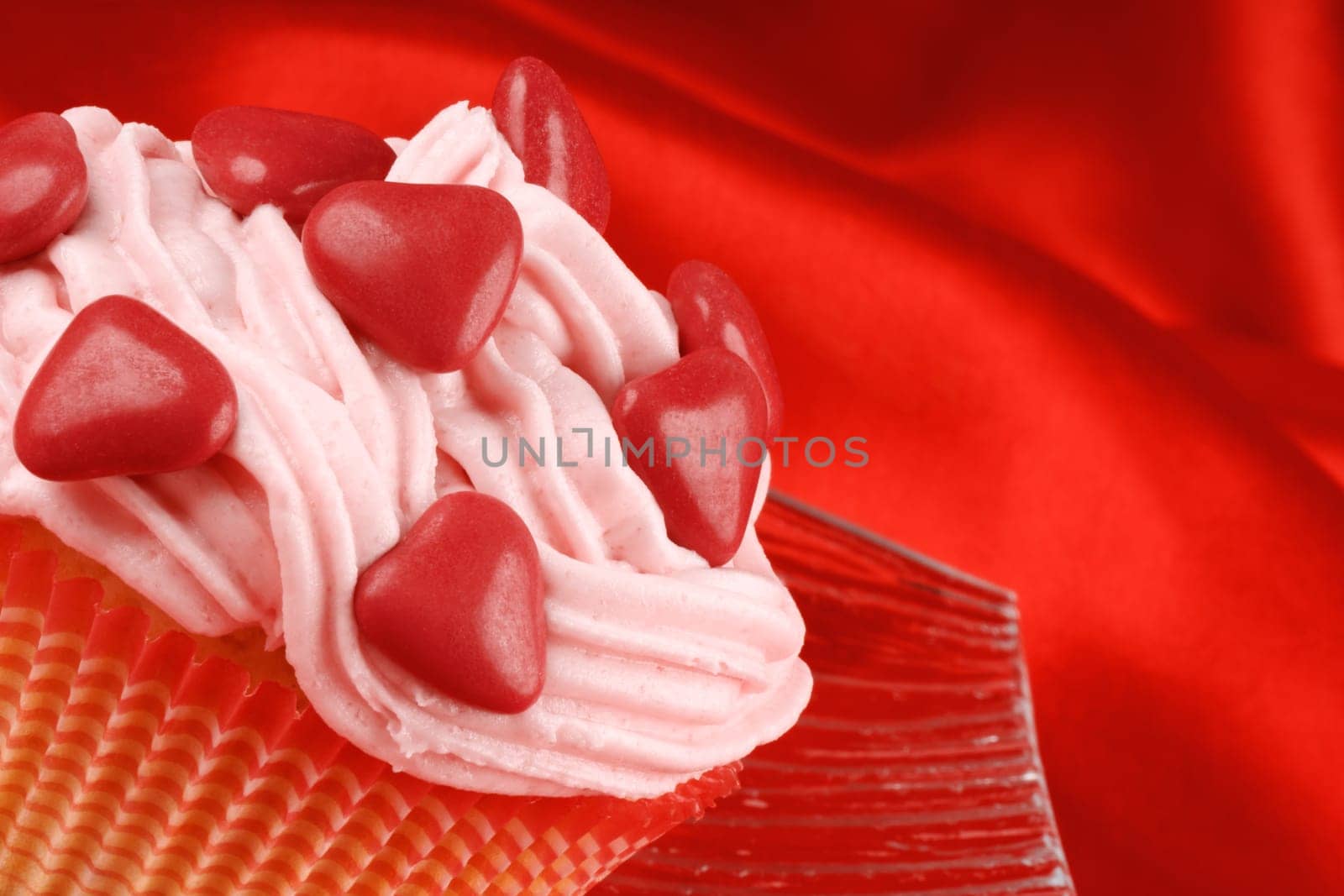 Fancy Valentine&#039 s Day cupcake decorated with small red sugar-coated chocolate hearts on a transparent glass dish, over a red background. Selective focus.