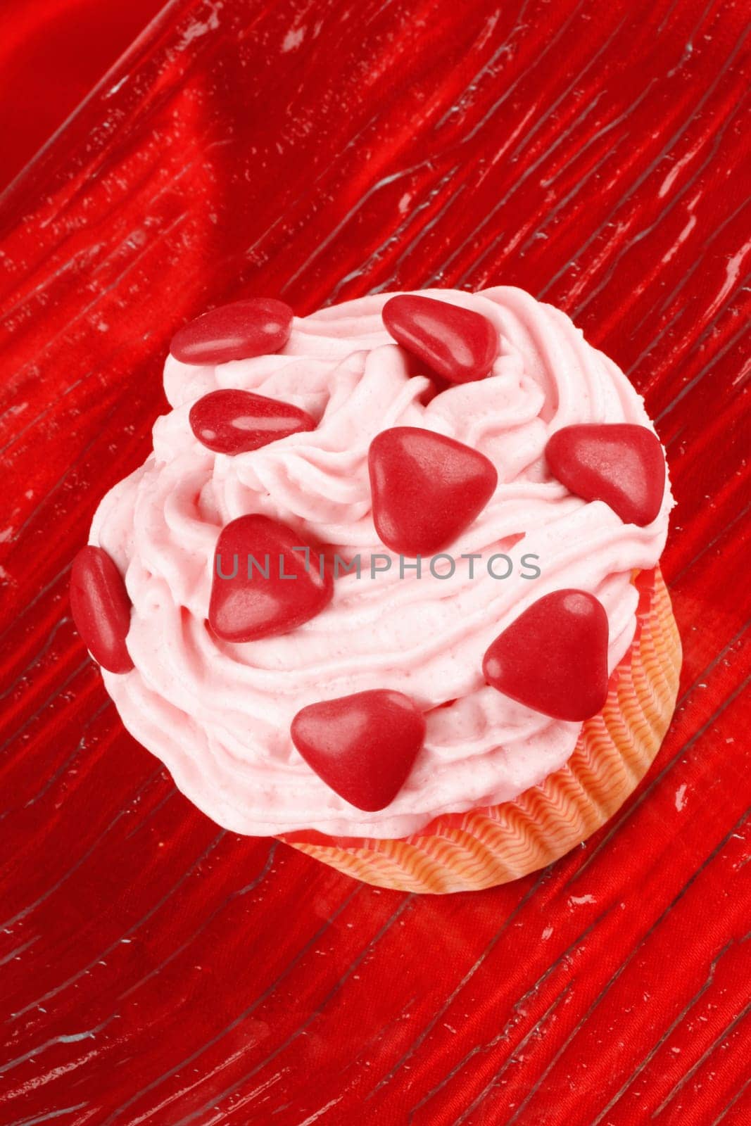 Fancy Valentine&#039 s Day cupcake decorated with small red sugar-coated chocolate hearts on a transparent glass dish, over a red background. Selective focus.