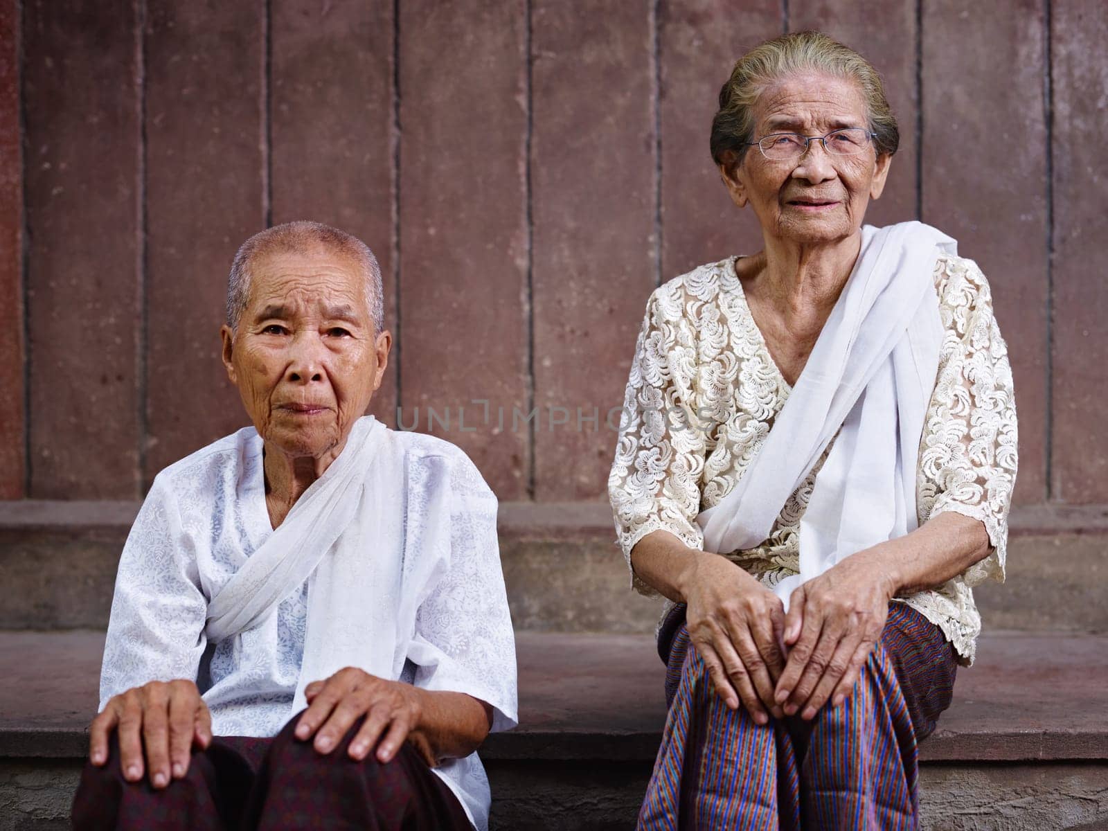 Portrait of two old asian women sitting against brown wall and looking at camera