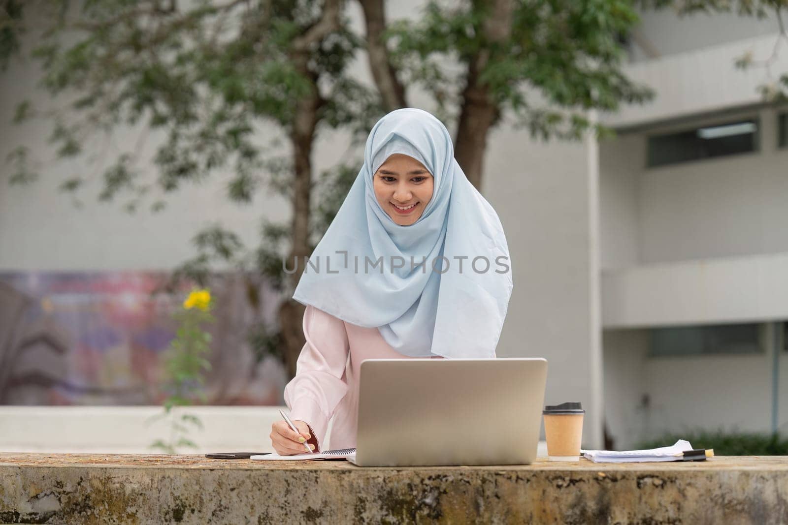 A smiling Muslim woman in a hijab studies with her laptop in a natural setting, representing her determination and growth in academics.