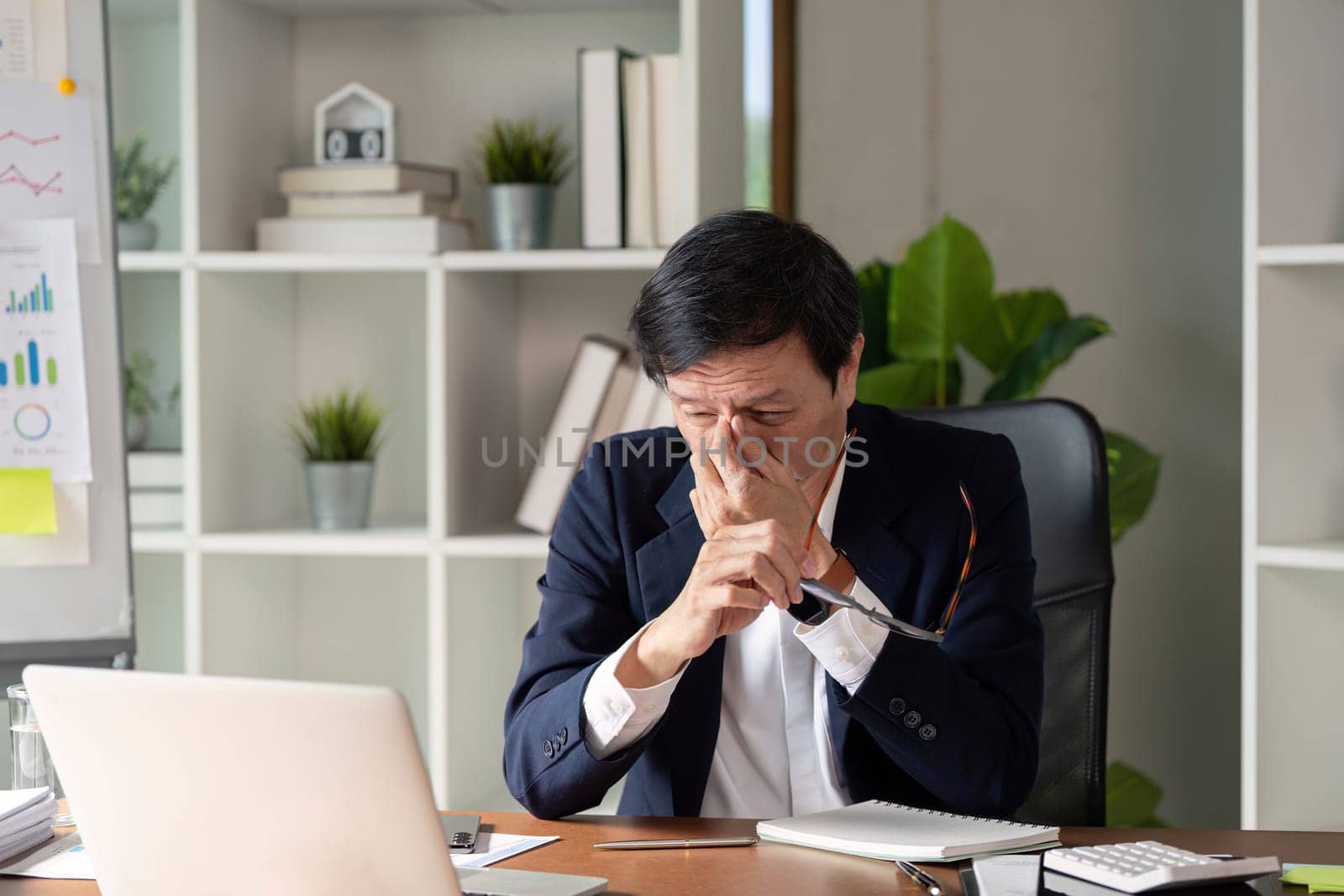 An elderly professional appears stressed while reviewing financial data on a laptop in a contemporary office space.
