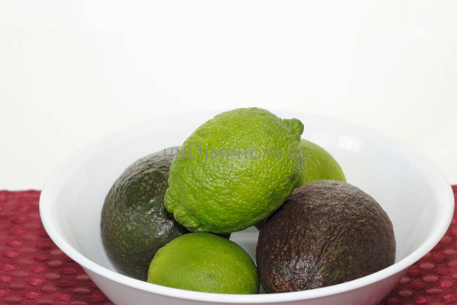Three raw limes and two avocados in a white bowl on a red place mat with an almost white background.