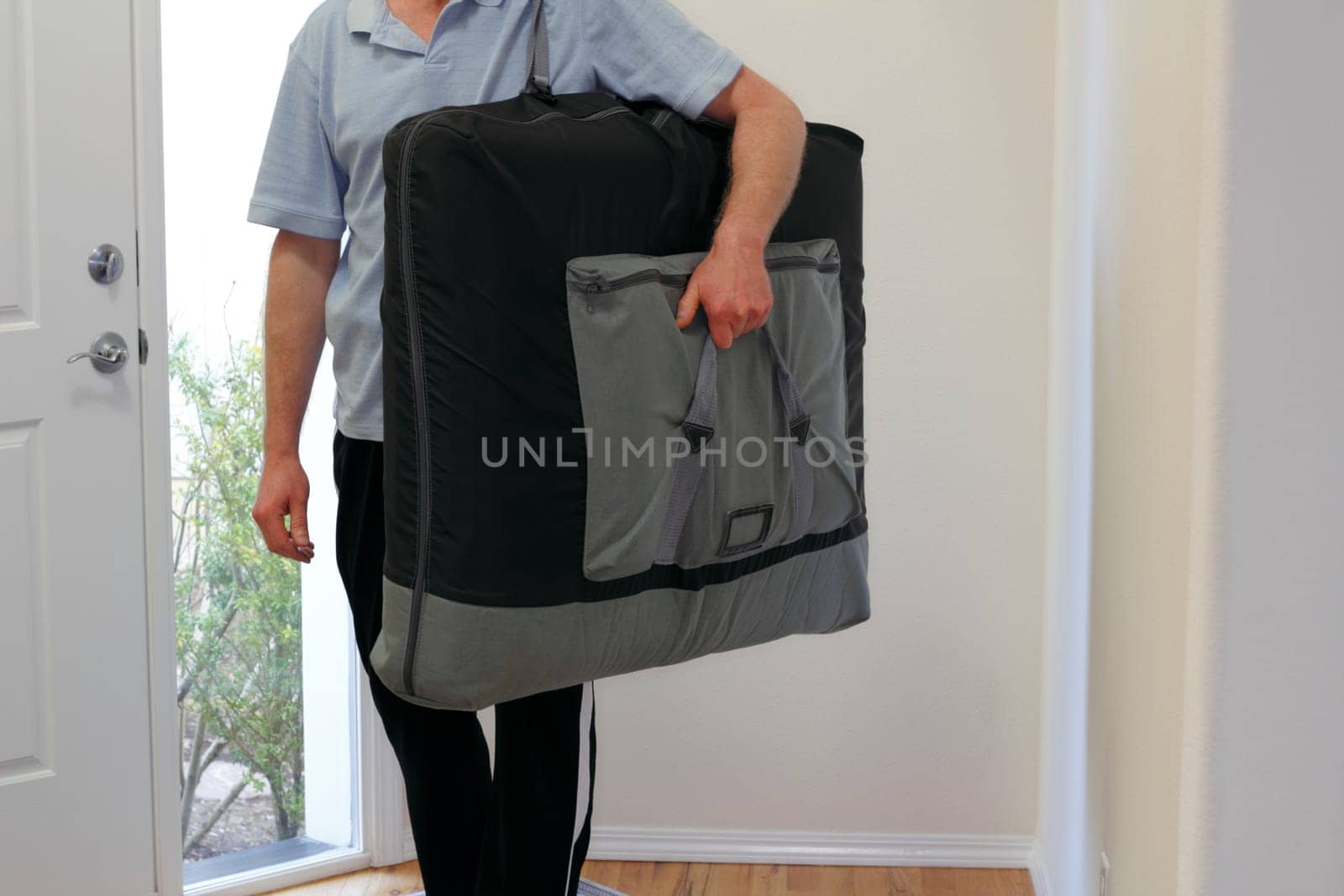 Standing inside the front door of a home a massage therapist is ready to set up the massage table he is carrying and massage his client.